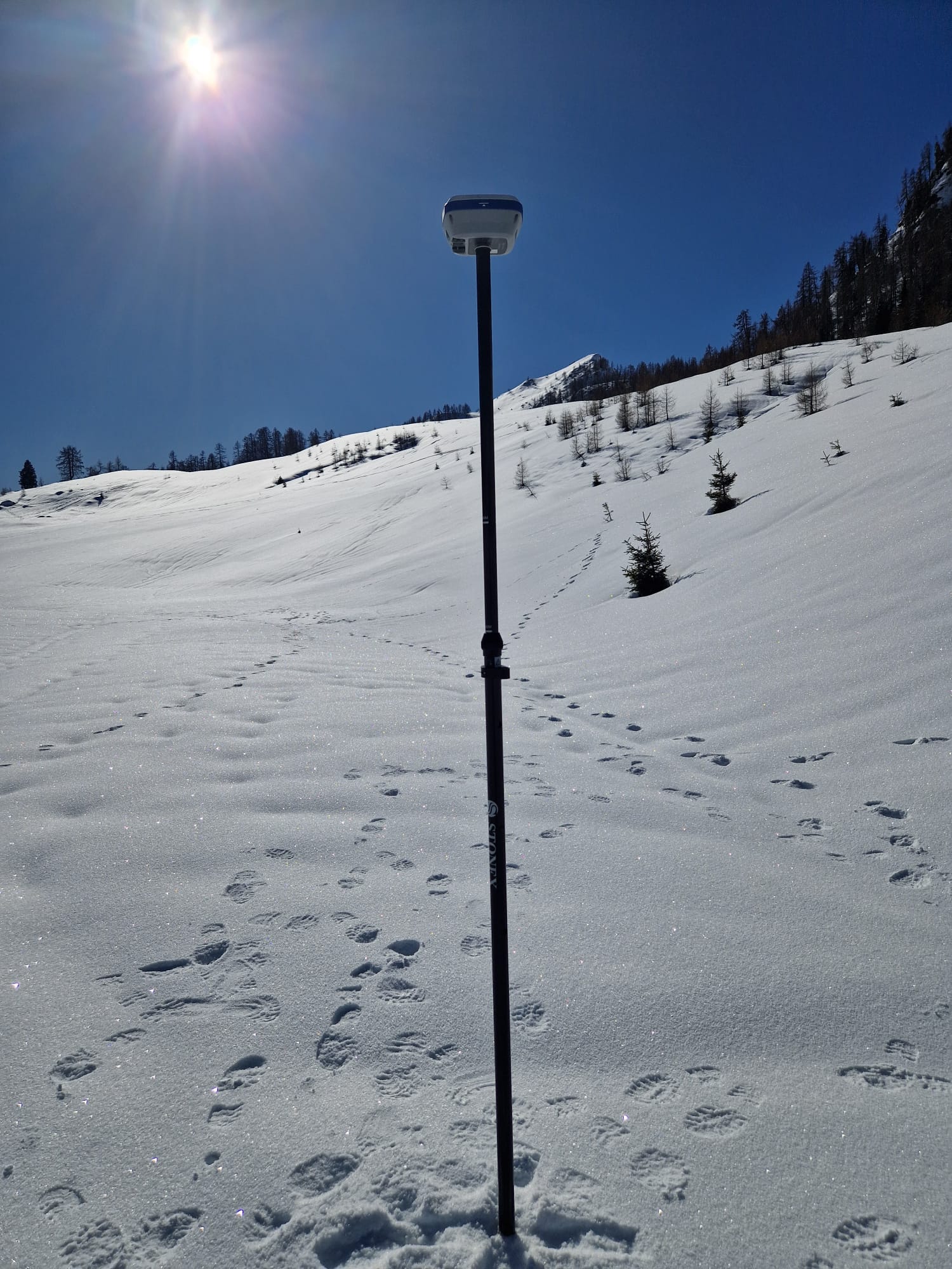 Rilievo GPS in ambiente innevato: antenna montata su palina al centro di un ampio pendio innevato sotto cielo azzurro, con il sole alto e tracce di passi sulla neve.